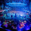 Širokoúhlá fotografie z pohledu za kapelou na koncertní scénu s davem diváků v sále. V popředí jsou vidět dvě bicí soupravy, keyboardy a muzikanti v…