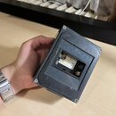 Černý plastový box s obdélníkovým výřezem, ve kterém je umístěna baterie s nápisem 9V. Povrch boxu vykazuje známky opotřebení a drobné znečištění. Bo…