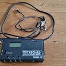 Černý napájecí adaptér s nápisem ROCKBOARD by WARWICK POWER PIT. Zařízení má digitální displej, přepínač napětí s nápisem Unplug pedal when selecting…