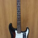Elektrická kytara s tmelem v barvě sunburst a bílým pickguardem. Na hlavě hředu je logo Fender a nápis STRATOCASTER, na pickguardu jsou viditelné náp…