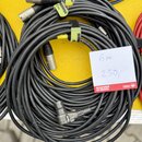 Svinutý černý kabel s konektory typu XLR na obou koncích. Kabel je stahován žlutými plastovými stěživkami a černým suchým zipem. Vedle kabelu je bílý…