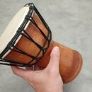 Dřevěný buben s bílou blánou, která je natažená a upevněna pomocí černého provázku. Tělo nástroje má hnědou barvu dřeva a zužující se tvar. Na povrch…