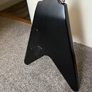 Zadní strana elektrické kytary s matným černým povrchem v tvaru V. Na zadní straně těla je vidět čtvercový otvor pro elektroniku zakrytý černým plátn…