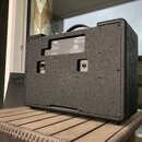 Zadní strana kytarového komba potaženého černým texturovaným povrchem. Na zadním panelu je vidět vstup pro napájení a ventilace. V horní části se nac…