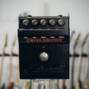 Černý kytarový efekt s nápisem DRIVE MASTER a logem Marshall v dolní části. Na horní straně jsou čtyři otočné knoflíky s oranžovými značkami, nad nim…