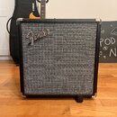 Černý kytarový kombo zesilovač s šedou pletenou přední síťovkou a kovovými rohy. V horní části sítě je umístěno stříbrné logo Fender. Nástroj stojí n…