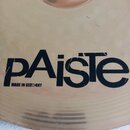 Detailní záběr na kovový činel v zlatavé barvě. Na povrchu je černý nápis pAiSte a pod ním menší text MADE IN GERMANY. Logo vykazuje známky opotřeben…