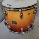Kytarový nebo bubnový nástroj, konkrétně tom, s povrchem v oranžovém gradientu (sunburst). Na bláně je viditelný nápis EVANS a na korpusu kulatá nále…