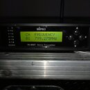 Černý přístroj s nápisem MiPro a označením M1-808T Stereo Transmitter. Vprostřed je umístěn žlutý displej s textem CH FREQUENCY 01 799.275MHz. Vlevo…