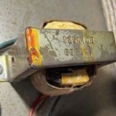 Fotografie zachicuje elektrický transformátor s kovovým jádrem a vinutím v papírovém obalu. Na kovovém povrchu je vyražen nápis 125A10B a pod ním čís…