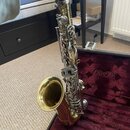 Zlatý saxofon umístěný v otevřeném kufru s burgundy vnitřkem. Nástroj má kovový povrch a stříbrné klapy. Na těle nástroje nejsou žádná čitelná loga a…