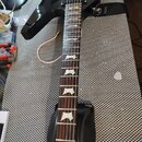 Černá elektrická kytara s hnědým hmatníkem a bílými vložkami v podobě trapézů. Na kytaru je nasazen černý popruh s nápisem ELIOTT GUITARS. Povrch kyt…