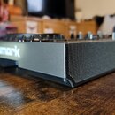 Černý DJ kontrolér s nápisem Numark na přední straně. Zařízení má hranatý tvar a v pravé části je vidět mřížka integrovaného reproduktoru. Povrch je…