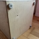 Světlá dřevěná bedna z překližky s viditelnou strukturou vrstev na hranách. V rozích jsou připevněny čtyři kulaté šedé filcové podložky. Na povrchu d…