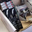 V kartonové krabici jsou dvě černé desky plošných spojů s bílými nápisy +5V, +12V a -12V u červených LED diod. Na deskách jsou bílé symboly blesku a…