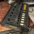 Černý kytarový syntetizér s nápisem KORG GUITAR SYNTHESIZER na spodní hraně. Na horním panelu jsou umístěny různé otočné potenciometry, přepínače a ž…