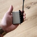 Stříbrný kovový přístroj obdélníkového tvaru s černou anténou. Na boku jsou vidět čtyři malé černé oválné prvky. Povrch je matný a bez viditelných ná…