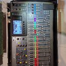 Digitální mixážní pult v černé barvě umístěný v transportním kufru. Na levé straně je vidět nápis X32 a v dolní části logo Behringer. Zařízení má bar…