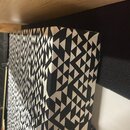 Detailní záběr na hranu a povrch akustického panelu. Panel je potažen látkou s černobílým geometrickým vzorem tvořeným trojúhelníky. Na fotografii ne…