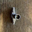 Stříbrná kovová ladicí mechanika s kolíkem. Povrch je lesklý, bez viditelných značek či nápisů. Předmět leží na dřevěném povrchu.