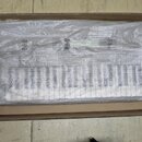 V kartonové krabici se nachází klaviatura zabalená v průhledné plastové fólii. Na fólii jsou vidět opakující se symboly recyklace a varovné piktogram…