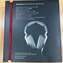 Fotografie zachycuje černý informační leták s technickými specifikacemi sluchátek. Na papíře je zobrazen pár šedých sluchátek s kovovými prvky a nápi…