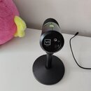 Černý mikrofon na kulatém stojanu. Na zadní straně je bílé logo VI a pod ním menší nečitelný nápis, dále jsou vidět konektory pro USB a jack. Horní č…