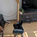 Černá elektrická kytara s přírodním dřevěným hřdelem a krčkem, umístěná na stojanu. Na hlavě je viditelné logo Squier. Na krytce elektroniky je nápis…