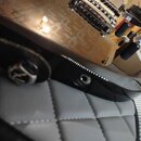 Detailní záběr na hranu těla elektrické kytary s hnědým povrchem a viditelnou strukturou dřeva. Na černé hraně těla je umístěn kovový popruhový háček…