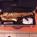 Zlatý saxofon v černém přepravním kufru. K nástroji je přiložen mouthpiece a bílé látkové sáčky. Na saxofonu nejsou viditelné žádné značky ani nápisy.