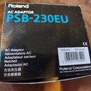 Černý kartonový obal od síťového adaptéru. Na horní části je bílým a modrým písmem nápis Roland AC ADAPTOR PSB-230EU. Na boční straně jsou uvedeny ná…