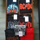 Fotografie zobrazuje sedm černých a jedno červené tričko s potisky kapely AC/DC. Každé tričko má připevněný bílý lístek s označením velikosti L nebo…
