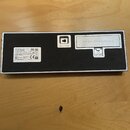 Zadní strana černého obdélníkového zařízení s kovovým rámem. Na povrchu je vypěstřovník s nápisem BATTERY: LR03 or 'AAA' SIZE x4, přepínač s označení…
