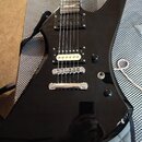 Černá elektrická kytara s ostrovním tvarem těla a hnědým hmatníkem. Nástroj má dva humbuckery, jeden ovládací knoflík a přepínač. Povrch je vysoce le…