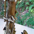 Detailní pohled na horní část mosazného saxofonu s viditelnou patinou a opotřebením povrchu. Nástroj je v matné zlatavé barvě s tmavšími skvrnami a š…
