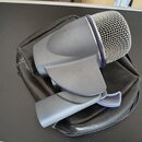 Šedý mikrofon s kovovou mřížkou, položený na černém koženém pouzdře. Na mikrofonu nejsou viditelné žádné značky ani nápisy. Povrch plastového těla vy…