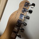 Zadní strana hlavy kytary z přírodního dřeva s šesti chromovanými ladičkami. Na každém ladičku je vyryto logo Fender. Na dřevě je černý nápis Model U…