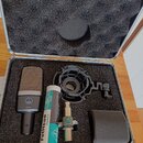 Otevřený hliníkový kufr s černé pěnovou výplní obsahuje kondenzátorový mikrofon značky AKG model C 214, šokabsorber a pop 필tr. Vedle mikrofonu je bíl…