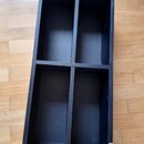 Černý dřevěný regál s čtyřmi otevřenými přihrádkami, umístěný na dřevěné podlaze. V pravém dolním rohu je vidět bílá nálepka s nápisem GLORIOUS. Povr…