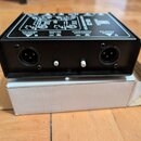 Černý kovový box s dvěma XLR konektory na zadní straně. Nad konektory jsou bílé nápisy BALANCED OUTPUT, GROUND GND LIFT a STEREO/MONO MONO STEREO. Me…