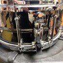 Černý snare drum s chromovaným kováním. Na mechanismu napnutí jsou viditelné nápisy "TAMA" a "SNARE". Nástroj vykazuje známky používání v podobě drob…