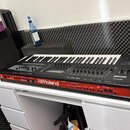 Černý klavír s červeným panelem v dolní části, umístěný na bílém stole. Na předním červeném panelu je bílý nápis Roland a menší text phantom. Horní o…