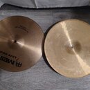 Dvě mosazné činely typu hi-hat položené vedle sebe na šedé podlaze. Levý činel nese nápisy MEINL, LIVE SOUND a 14" MEDIUM HI-HAT BOTTOM. Pravý činel…