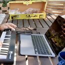 Na dřevěném stole je černý kompaktní klaviatura s bílými a černými klávesami, na které je nápis m-mini 32. Nástroj je připojen k stříbrnému notebooku…