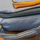 Detailní záběr na šedý textilní obal s oranžovými prvky. Je vidět černý zip a šedá rukojeť s oranžovým detailem. Na materiálu nejsou viditelné žádné…