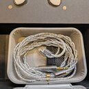 V šedém pouzdře je svinutý stříbrný pletený kabel. Na konektoru jacku je viditelný nápis "Symphonium". V horní části černého boxu jsou vidět části dv…