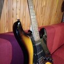 Elektrická kytara s tělem v žlutě-černé barvě (sunburst) a černým pickguardem. Nástroj má hриф z tmavého dřeva s viditelnými pražci. Na těle jsou tři…