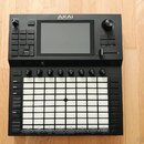 Černý MIDI kontrolér s bílou mřížkou 8x8 gumových padů. Na horní části je umístěn displej a nad ním nápis AKAI PROFESSIONAL. Zařízení obsahuje několi…