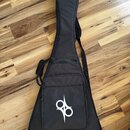 Černý polstrovatý pouzdro na kytaru s ramenním popruhem. Na přední kapse je bílé grafické logo připomínající stylizované písmeno S nebo symbol nekone…