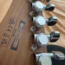 Detail hlavy hnědé dřevěné kytary s šesti chromovanými ladičkami. Na zadní straně hlavy je černý štítek s nápisy MADE IN CHINA, 5N250700760 a několik…